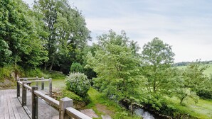 A wooden deck with a gravel path leading to a river, surrounded by lush greenery and trees. - 4 bedroom accommodation in Great Strickland near Ullswater (Great Strickland near Penrith)
