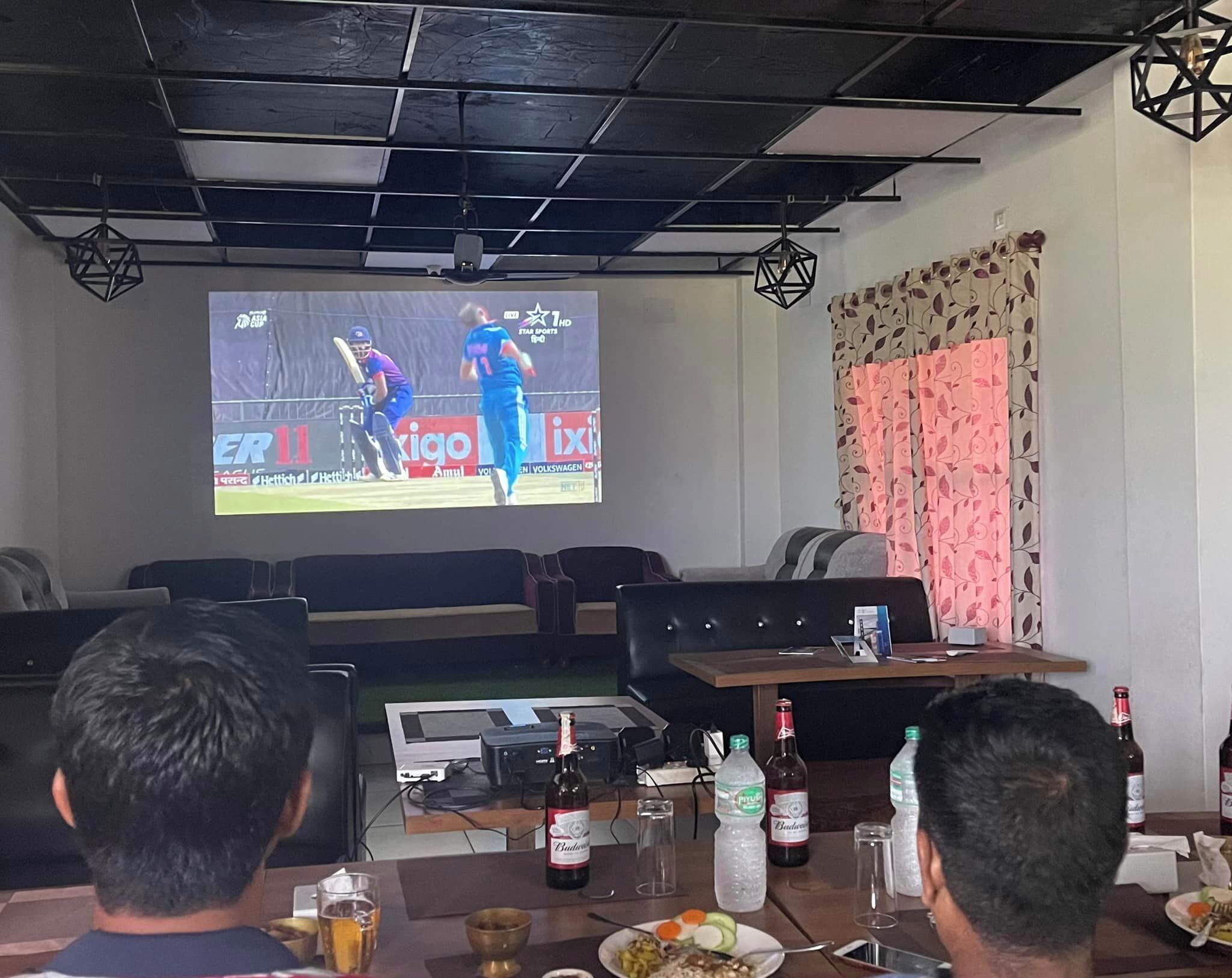 Una habitación con una gran pantalla mostrando un partido de criquet, una mesa con bebidas y aperitivos, y un proyector.