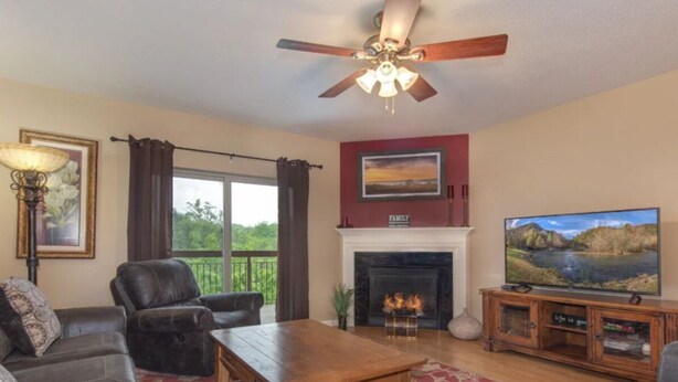 A living room with a fireplace, a ceiling fan, a television, and a large window with curtains. - Top-Floor Riverfront – 3BR Condo with Elevator Access & Double Vanities (Pigeon Forge)