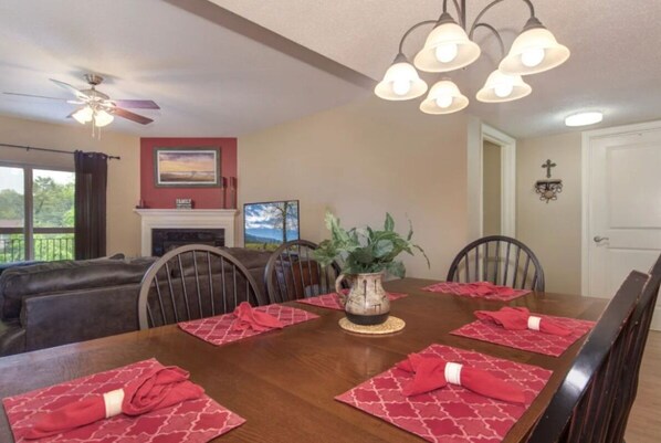A dining room with a wooden table set for four, red placemats, a potted plant, and a fireplace.