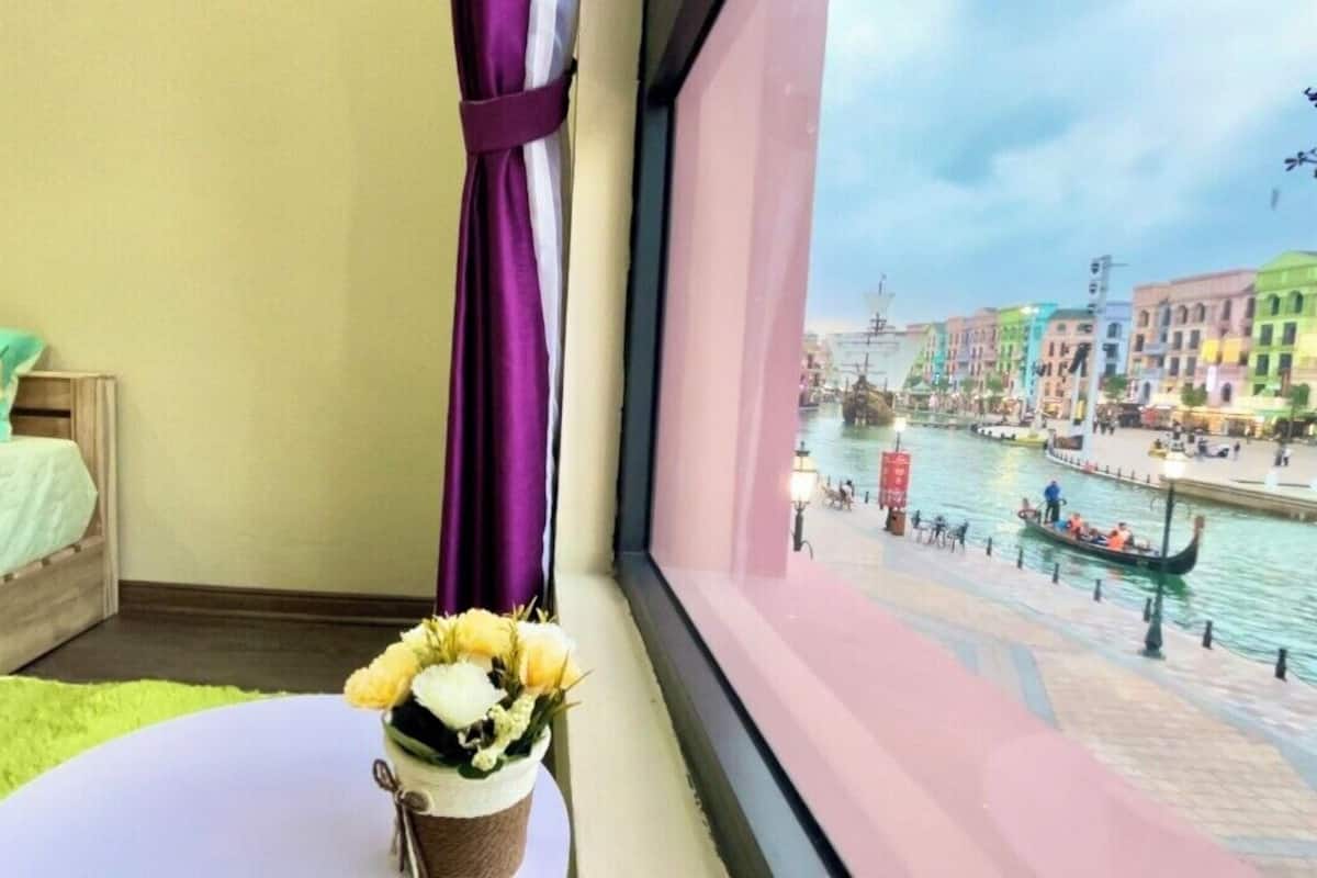 A room with a bed, a small table with a vase of flowers, and a window overlooking a canal with colorful buildings and a gondola.