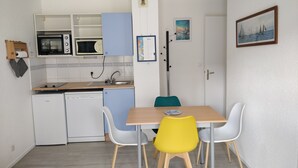 Kompakti keittiö, jossa on valkoiset kodinkoneet, ruokapöytä kahdella tuolilla ja pieni jääkaappi.