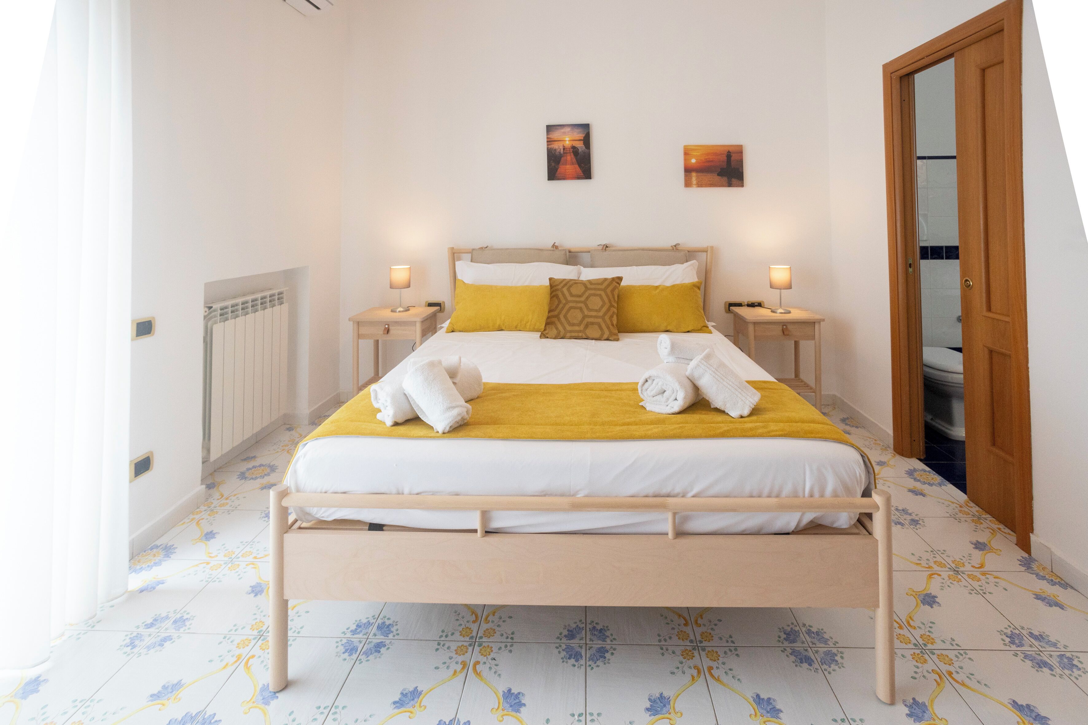 Un lit bien fait, avec une literie blanche et jaune, deux tables de chevet avec des lampes et un tapis au motif floral.