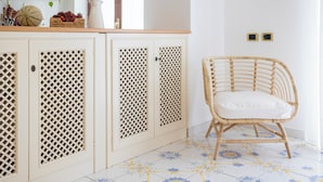 A kitchen with a white cabinet featuring a lattice design, a wicker chair with a white cushion, and a tiled floor with a floral pattern.