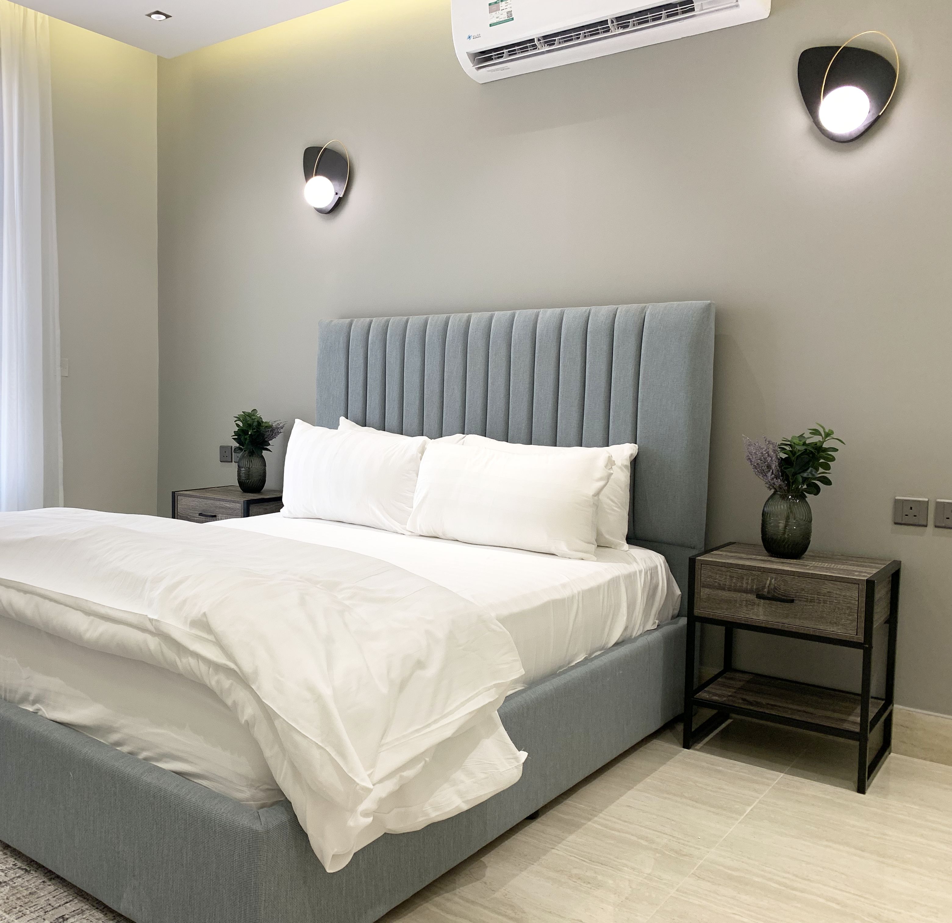 Un dormitorio moderno con una cama tapizada en gris, mesitas de noche con lámparas y un ventanal con cortinas de gasa.