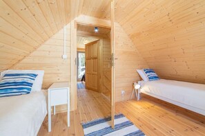 Tunnelmallinen ullakkohuone, jossa on puuseinät ja kaltevat katot. Huoneessa on kaksi sängyä, joissa on raidalliset tyynyt, pieni yöpöytä ja ovi, joka johtaa toiseen huoneeseen.