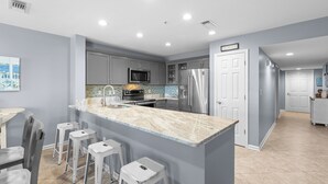 A modern kitchen with grey cabinets, stainless steel appliances, and a marble island with bar stools. - Amalfi Coast 108A- Community Pool, Great Amenities, Steps to Beach! (Miramar Beach)