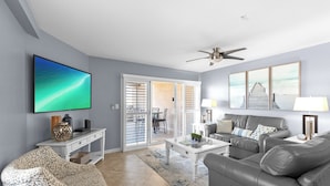 A living room with a flat-screen TV, a ceiling fan, a sofa, a coffee table, and a dining area with a sliding glass door. - Amalfi Coast 108A- Community Pool, Great Amenities, Steps to Beach! (Miramar Beach)