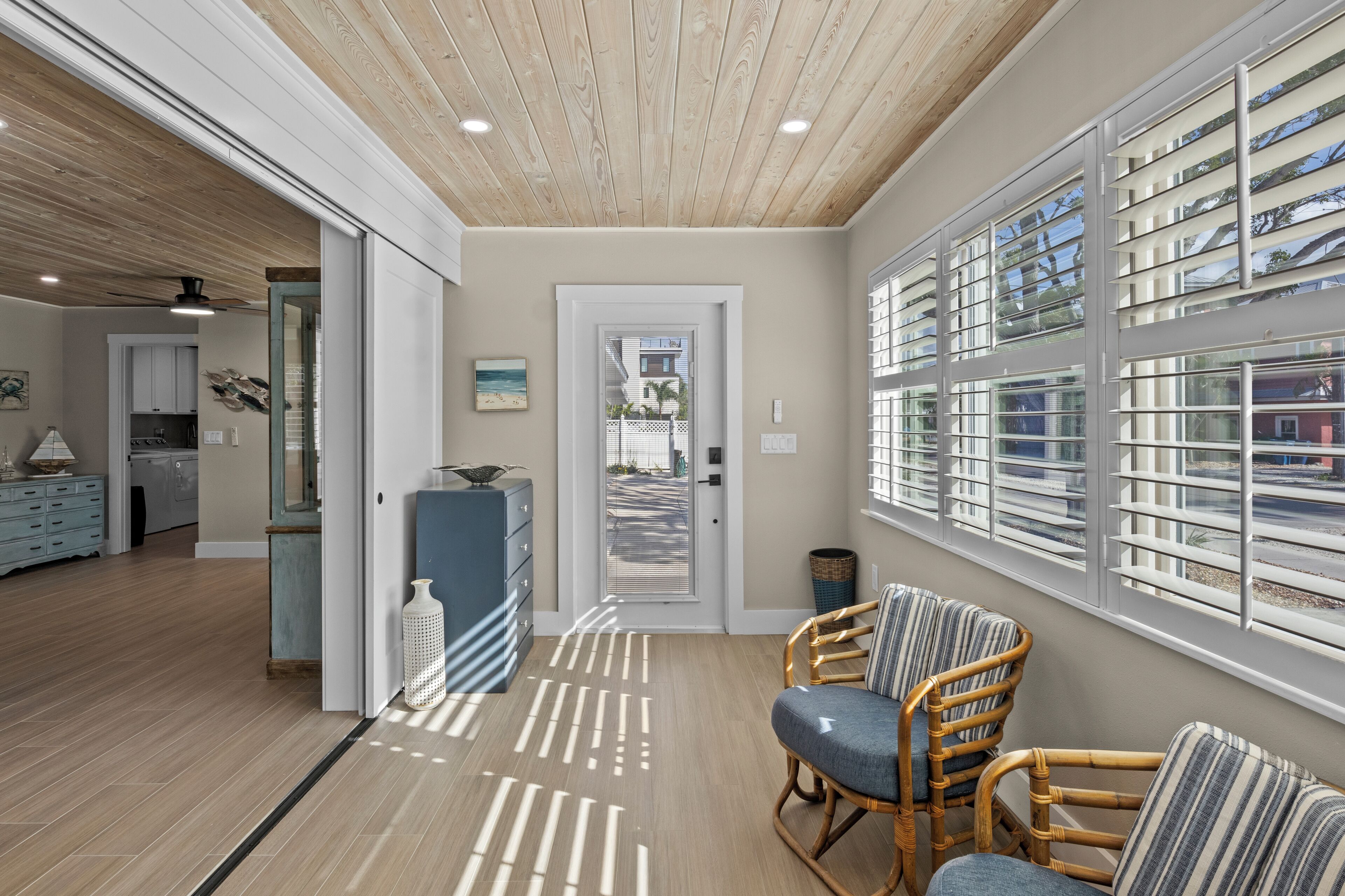 A modern interior with a wooden ceiling, a kitchen in the background, and a seating area with wicker chairs and a view through large windows.