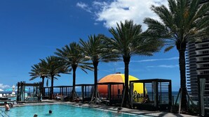 A swimming pool area with palm trees, a large sun-shaped structure, and a view of the ocean. - Lyfe Resort and residences, Ocean Front 35th floor, great views guaranteed! (Hollywood)