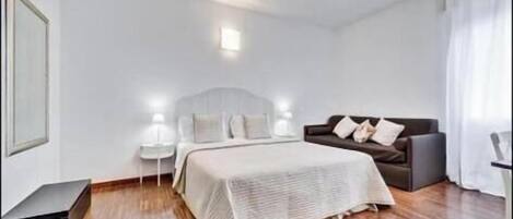 A bedroom with a bed, a sofa, a nightstand, and a wall-mounted light fixture.