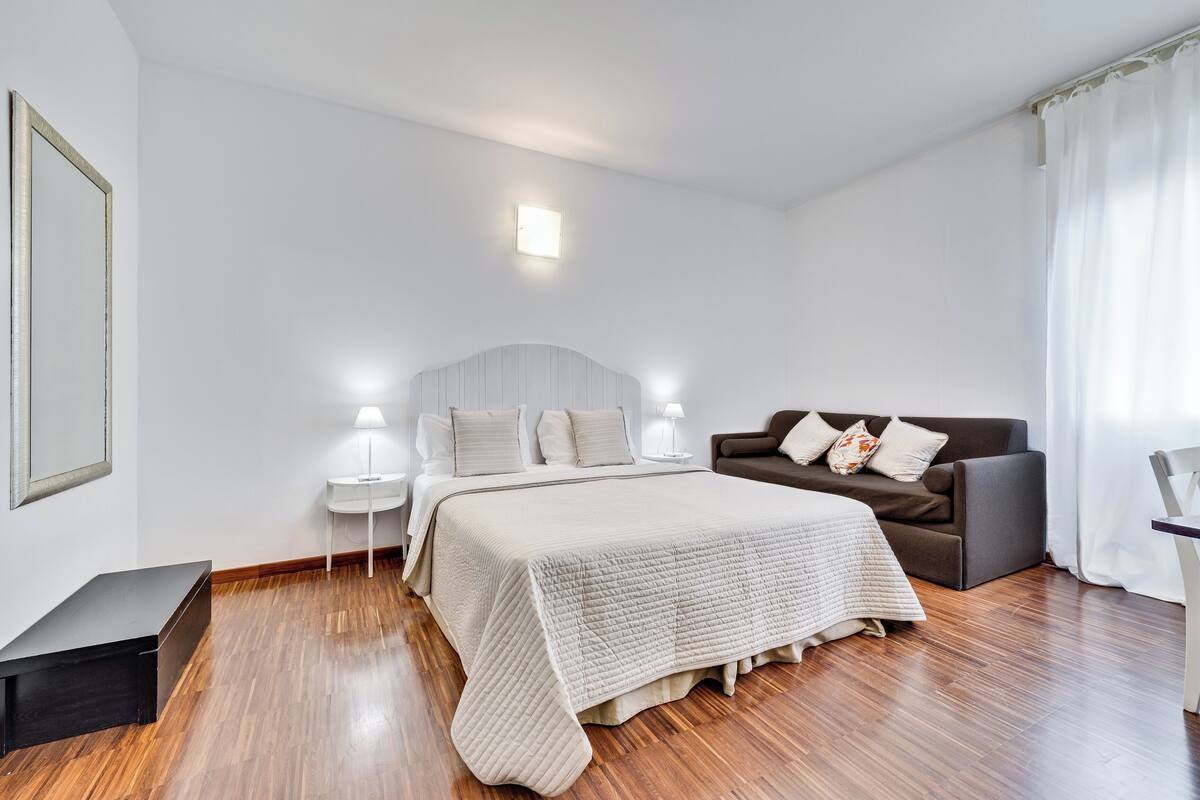 A bedroom with a bed, a nightstand, a lamp, a sofa, and a small table.