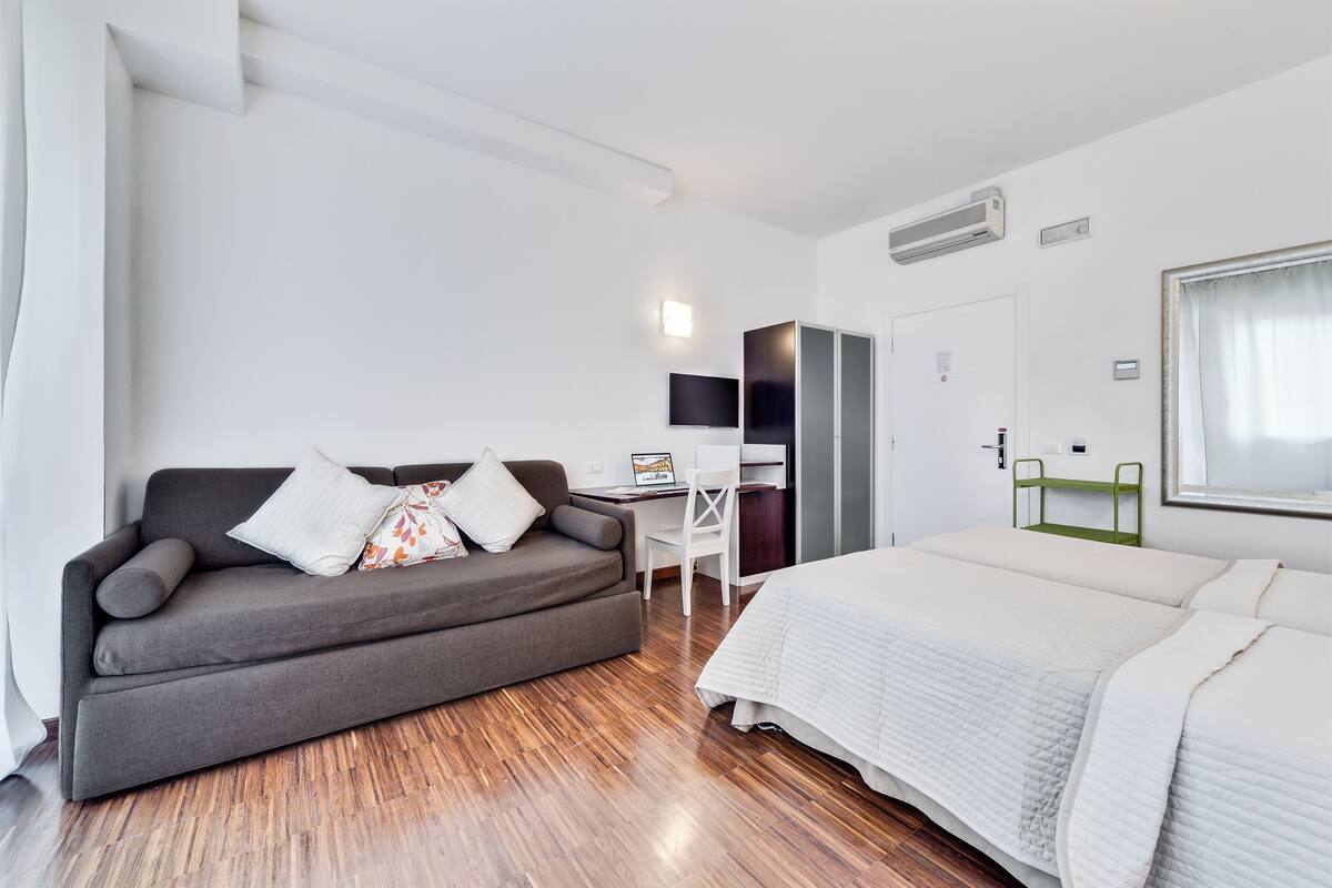 A modern hotel room with a grey sofa, a white bed, a desk with a chair, and a TV on the wall.