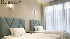 Hotelkamer met twee bedden, elk met een gestoffeerd hoofdeinde, witte beddengoed en een groot raam met dunne gordijnen.