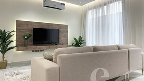 Een moderne woonkamer met een grote flatscreen-tv aan een houten wand, een beige bank en een groot raam met lichte gordijnen.