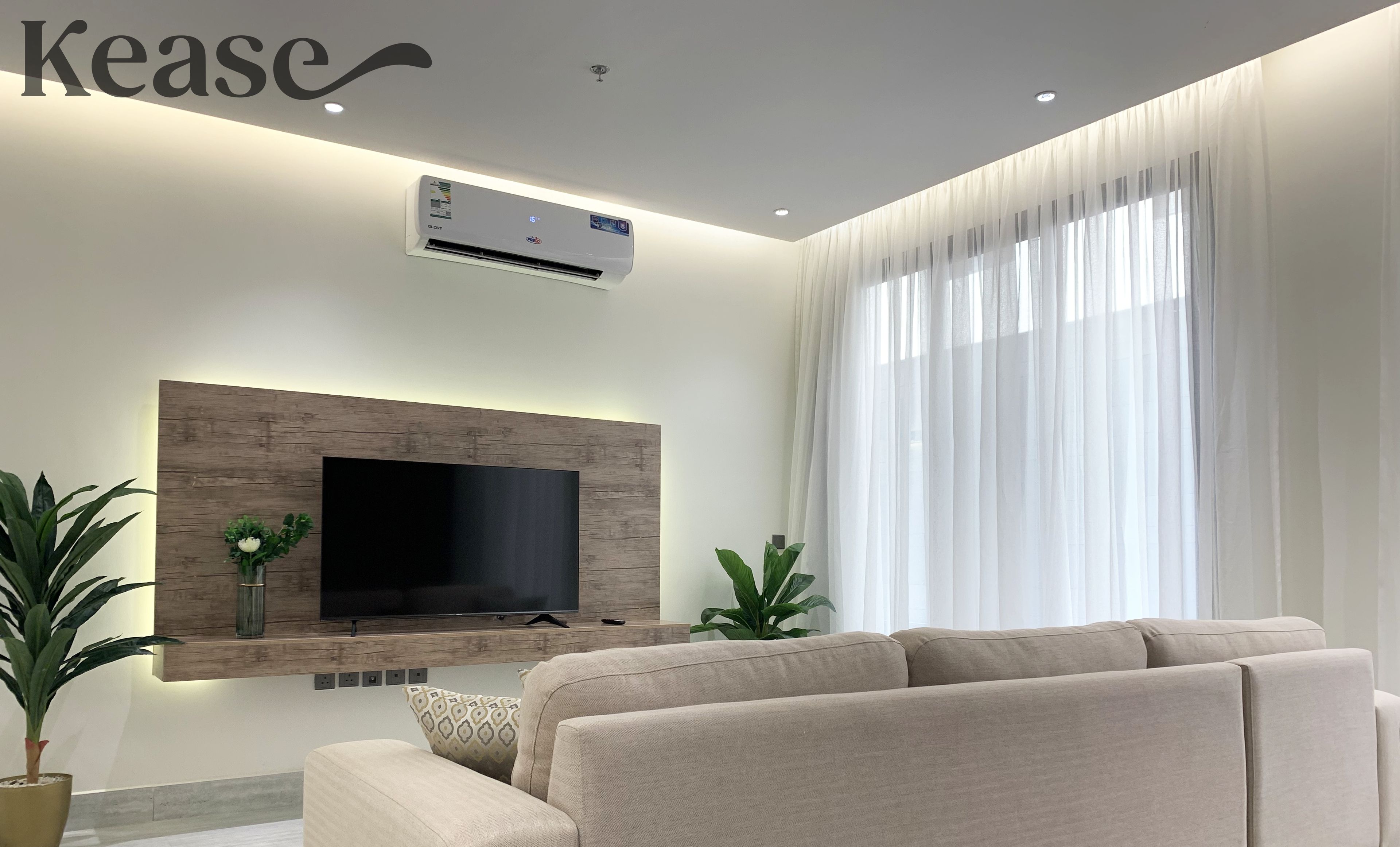 A modern living room with a large flat-screen TV mounted on a wooden wall, a beige sofa, and a large window with sheer curtains.