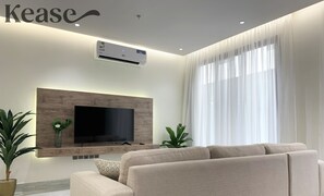 Un salon moderne doté d’un grand téléviseur à écran plat fixé sur un mur en bois, d’un canapé beige et d’une grande fenêtre ornée de rideaux légers.