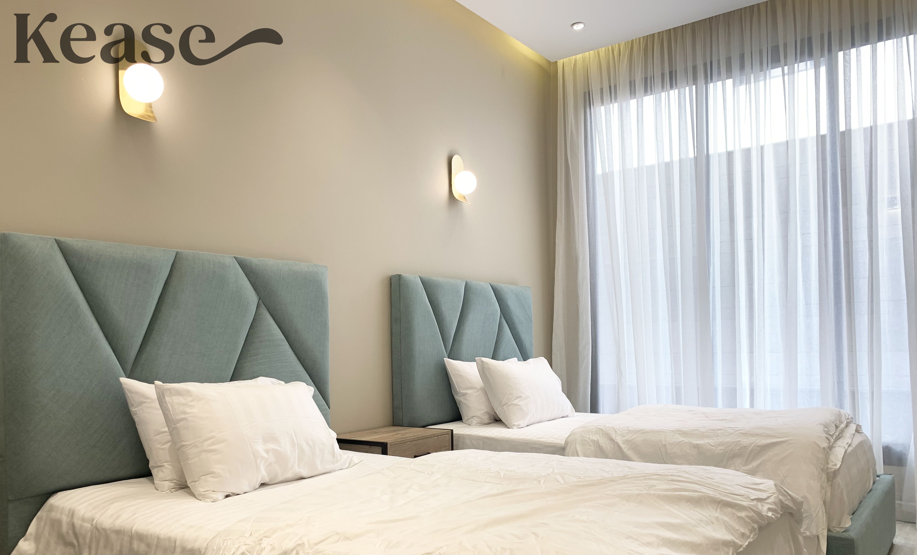 Hotellihuone, jossa on kaksi sänkyä. Jokaisessa sängyssä on koristeltu pääty, valkoiset vuodevaatteet ja suuri ikkuna, jossa on läpikuultavat verhot.