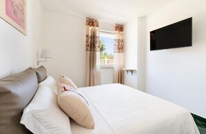 A neatly made bed with white linens and beige pillows, a flat-screen TV mounted on the wall, and a window with floral curtains.