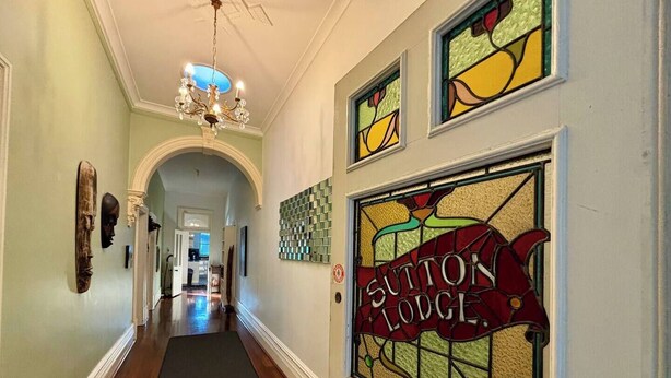 A hallway with a chandelier, a stained glass sign reading "Sutton Lodge," and a wooden headboard. - Subiaco Heritage House "Gem"! (Shenton Park)