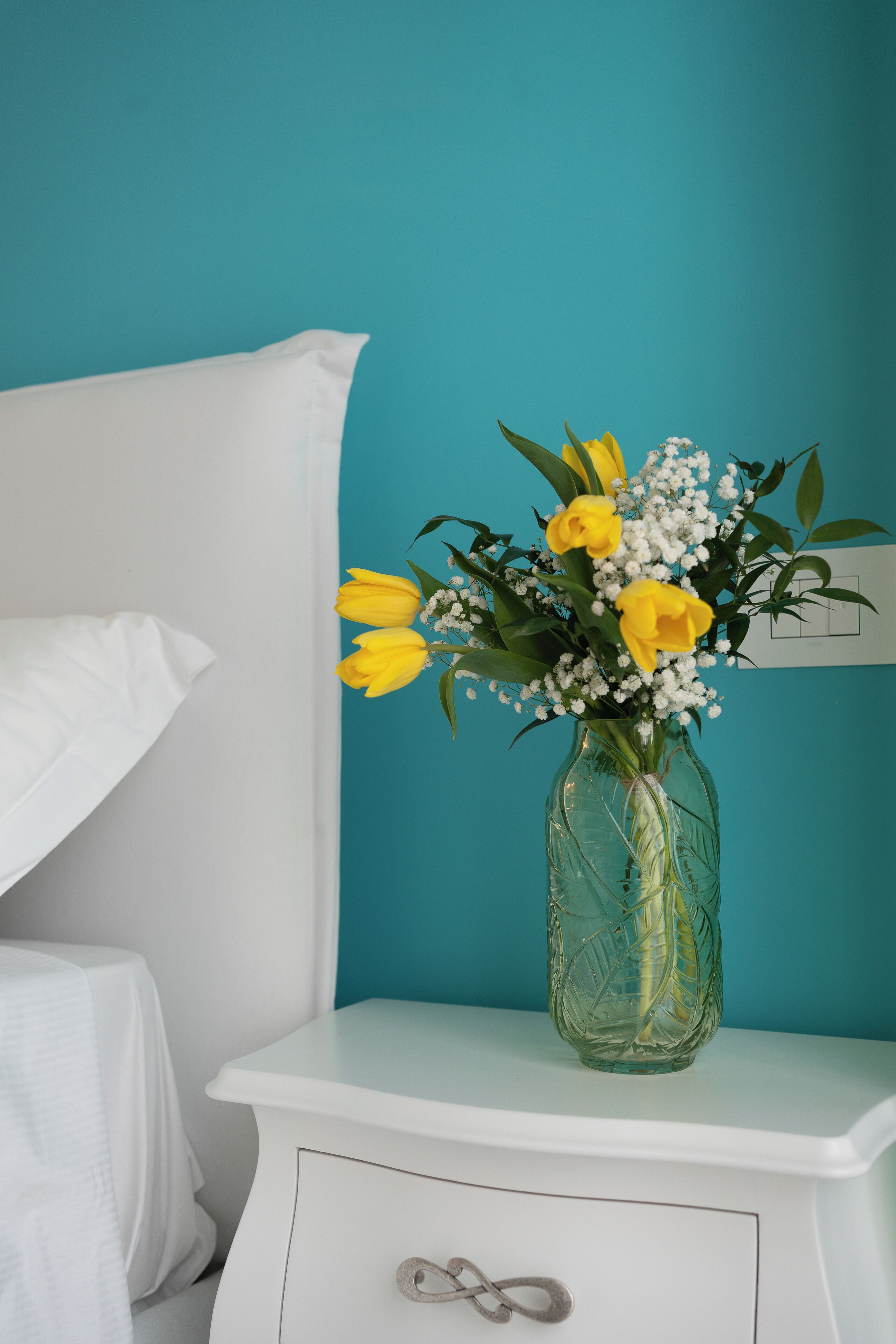 A white bedside table with a clear glass vase containing yellow flowers and white baby's breath against a teal wall.