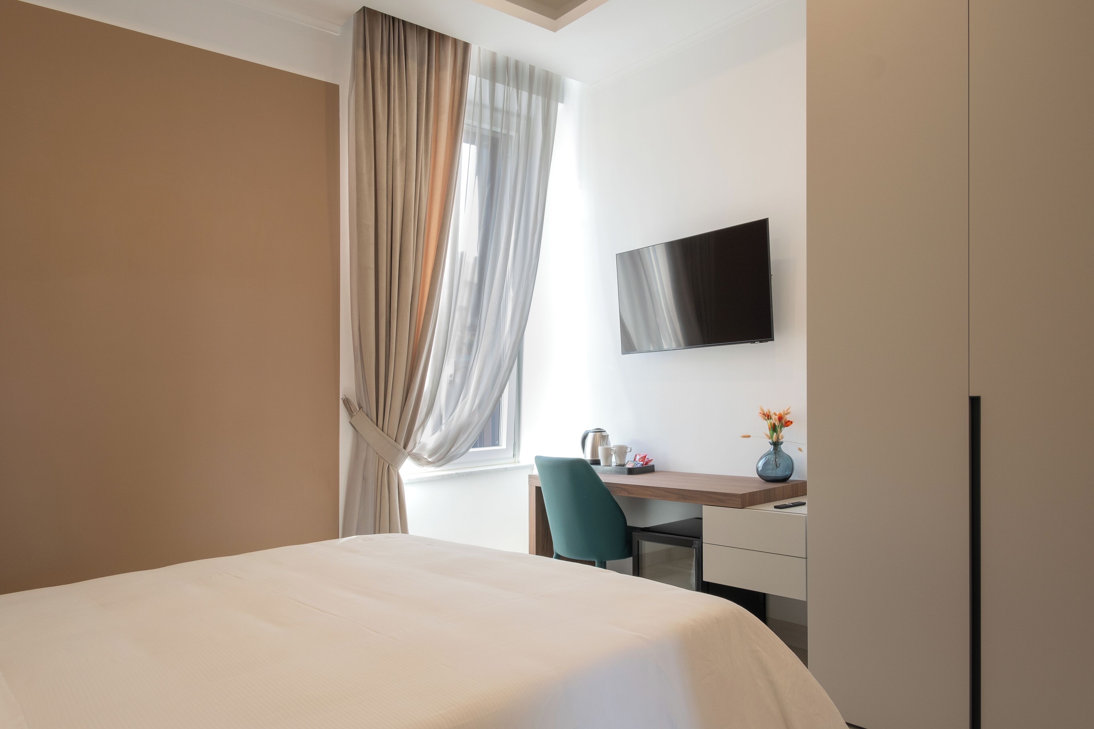 A hotel room with a bed, a desk with a chair, a TV mounted on the wall, and a window with curtains.