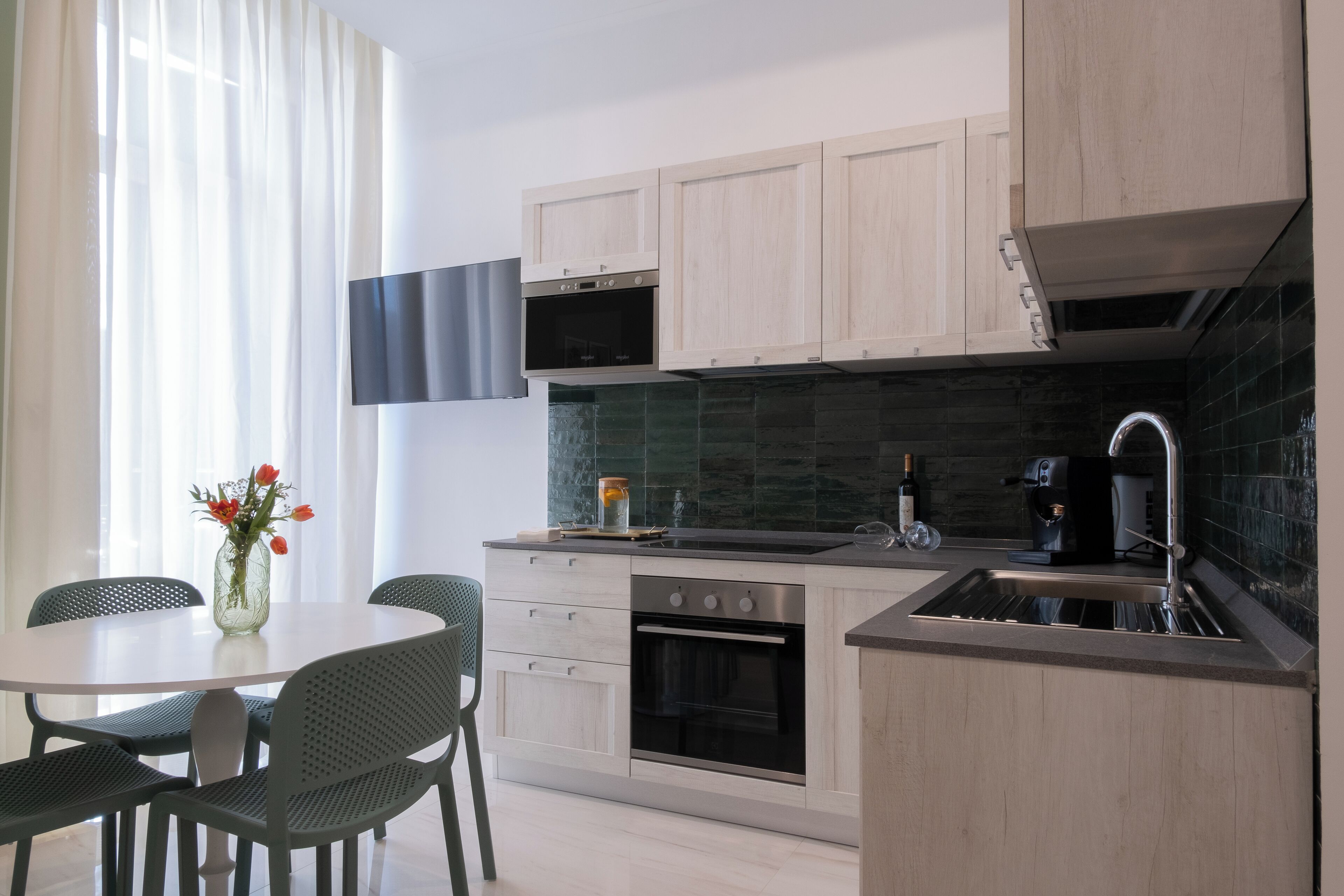Una cocina moderna con armarios de madera, un horno negro, un microondas y un lavamanos. Hay una mesa de comedor con sillas y un jarrón con flores.