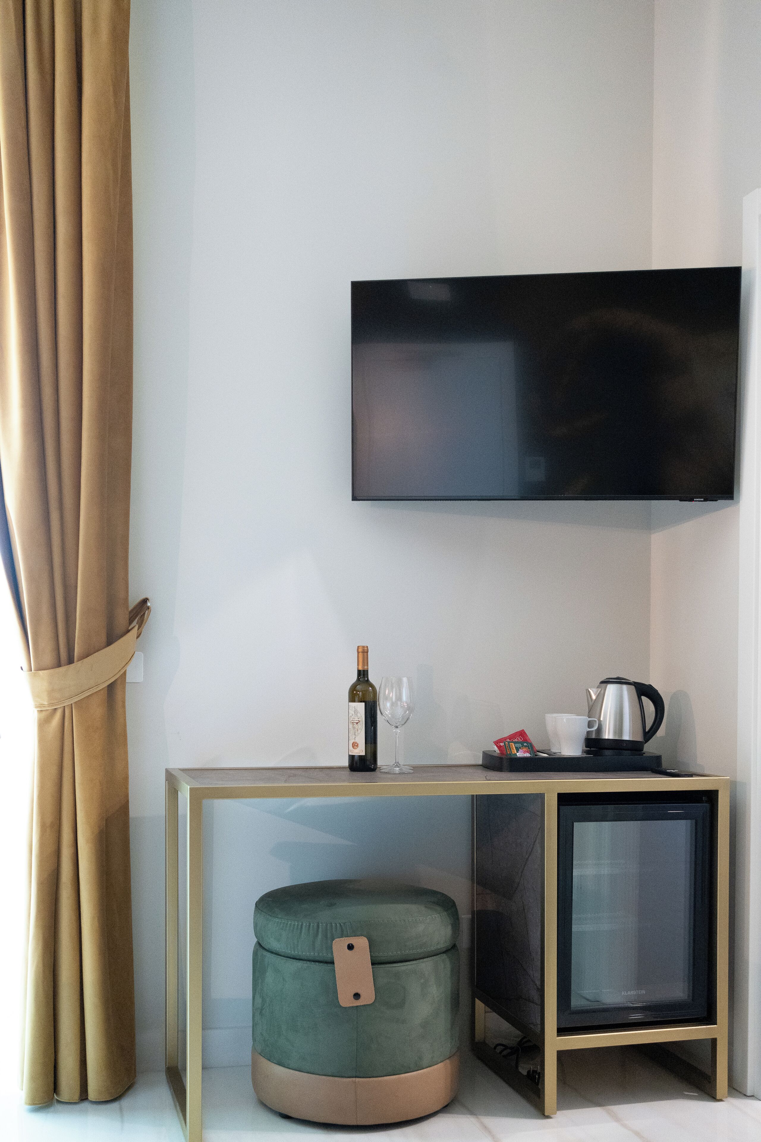 A modern room with a flat-screen TV mounted on the wall, a small cabinet with a kettle, and a cushioned ottoman.