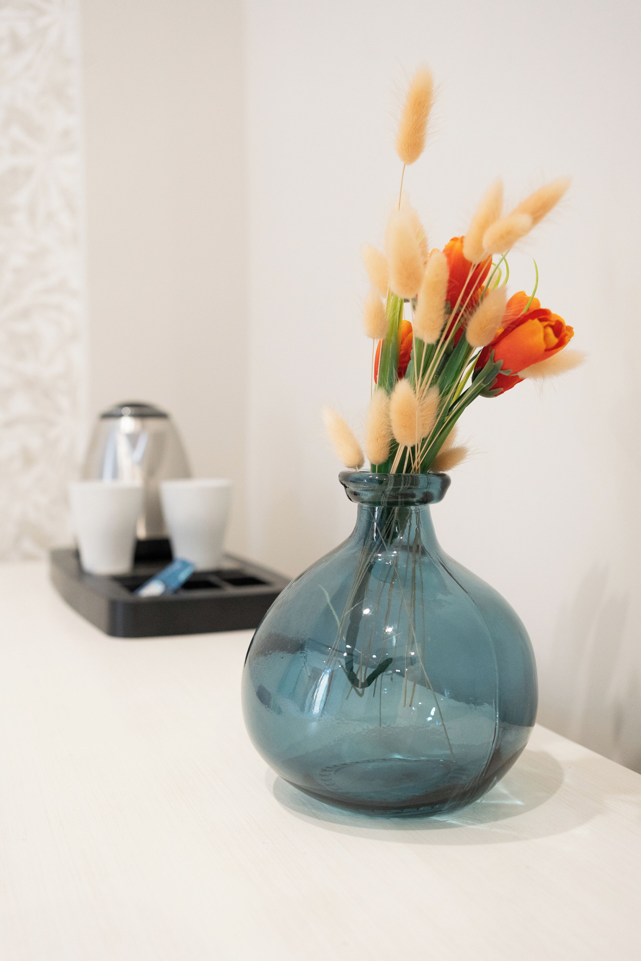 A clear glass vase with orange flowers and pampas grass on a white surface.