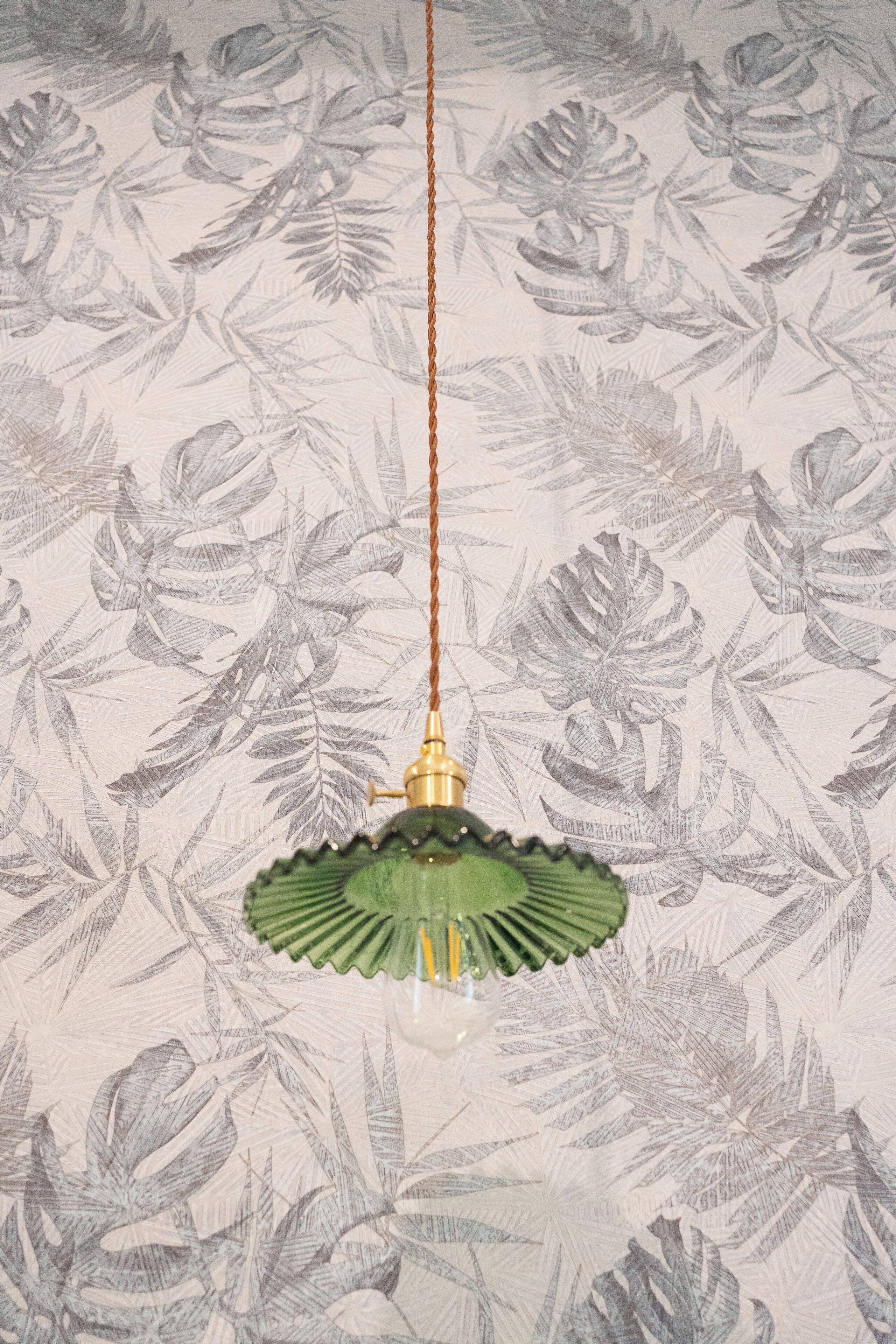 A hanging light fixture with a green shade and a brass base, suspended from a brown cord against a wallpaper with a leaf pattern.