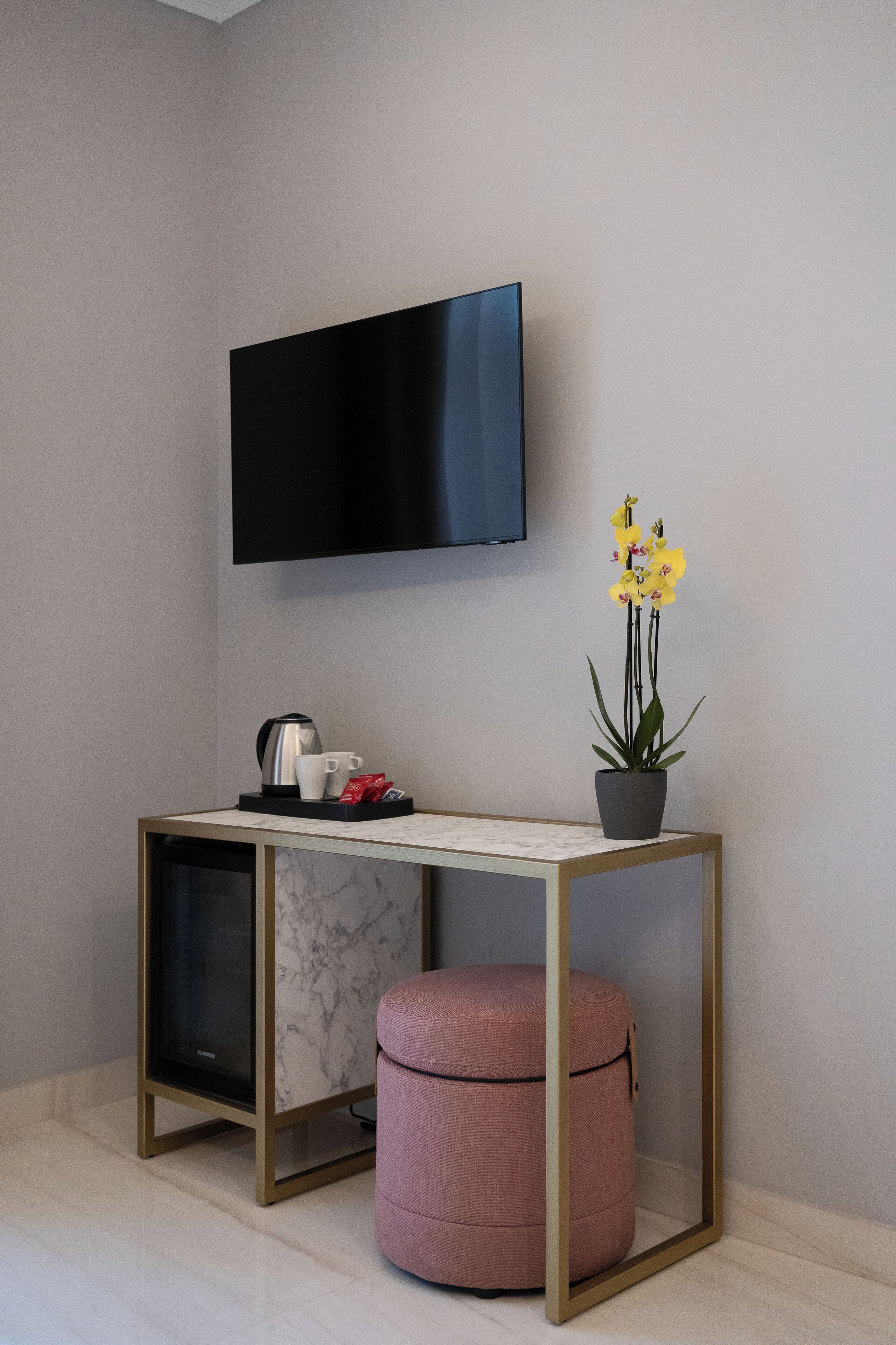 A modern room with a flat-screen TV mounted on the wall, a small table with a coffee maker, cups, and a potted plant.