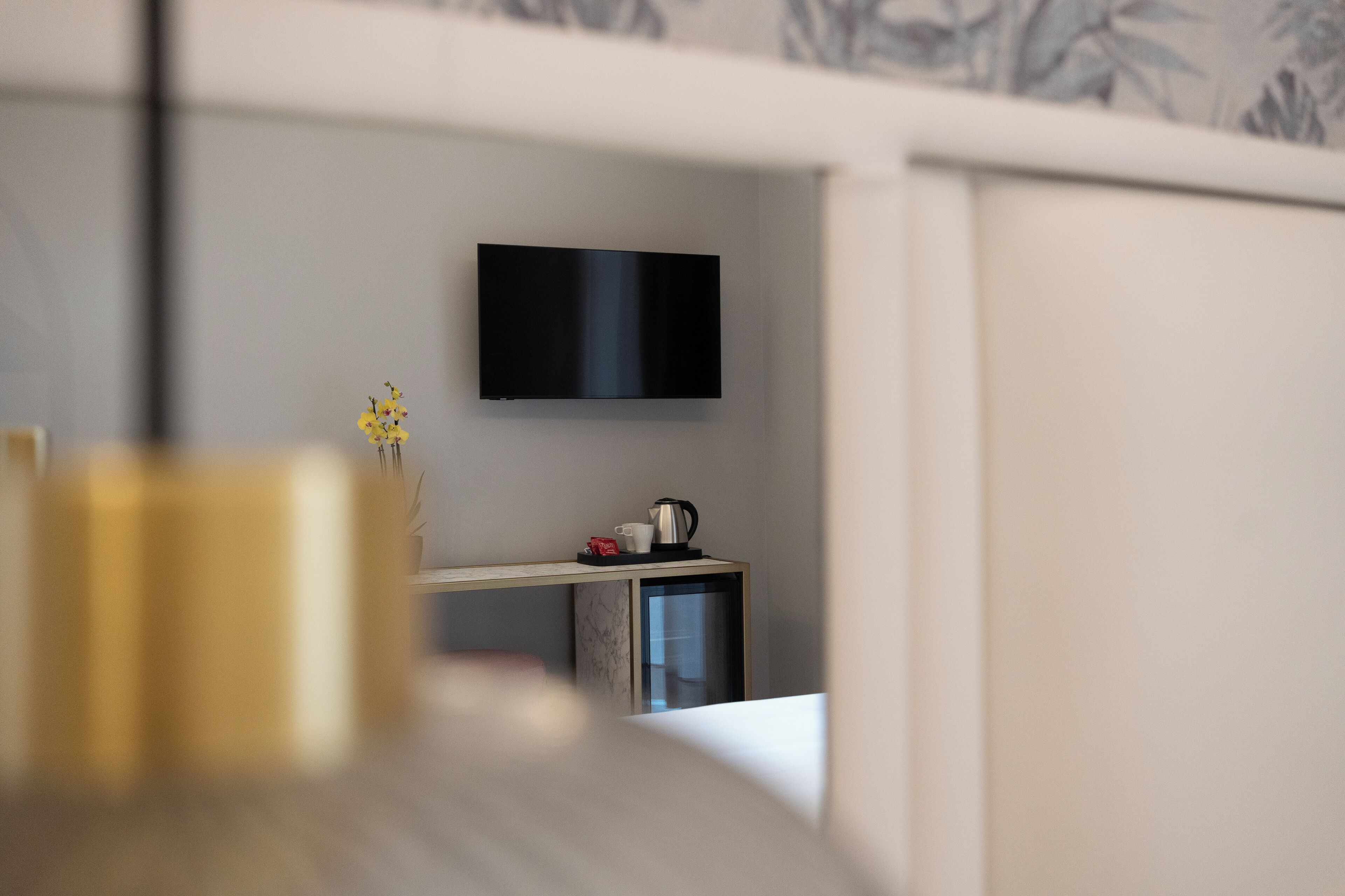 A modern hotel room with a flat-screen TV mounted on the wall, a small desk with a kettle and cups, and a vase with yellow flowers on the table.