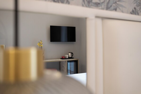 A modern hotel room with a flat-screen TV mounted on the wall, a small desk with a kettle and cups, and a vase with yellow flowers on the table.