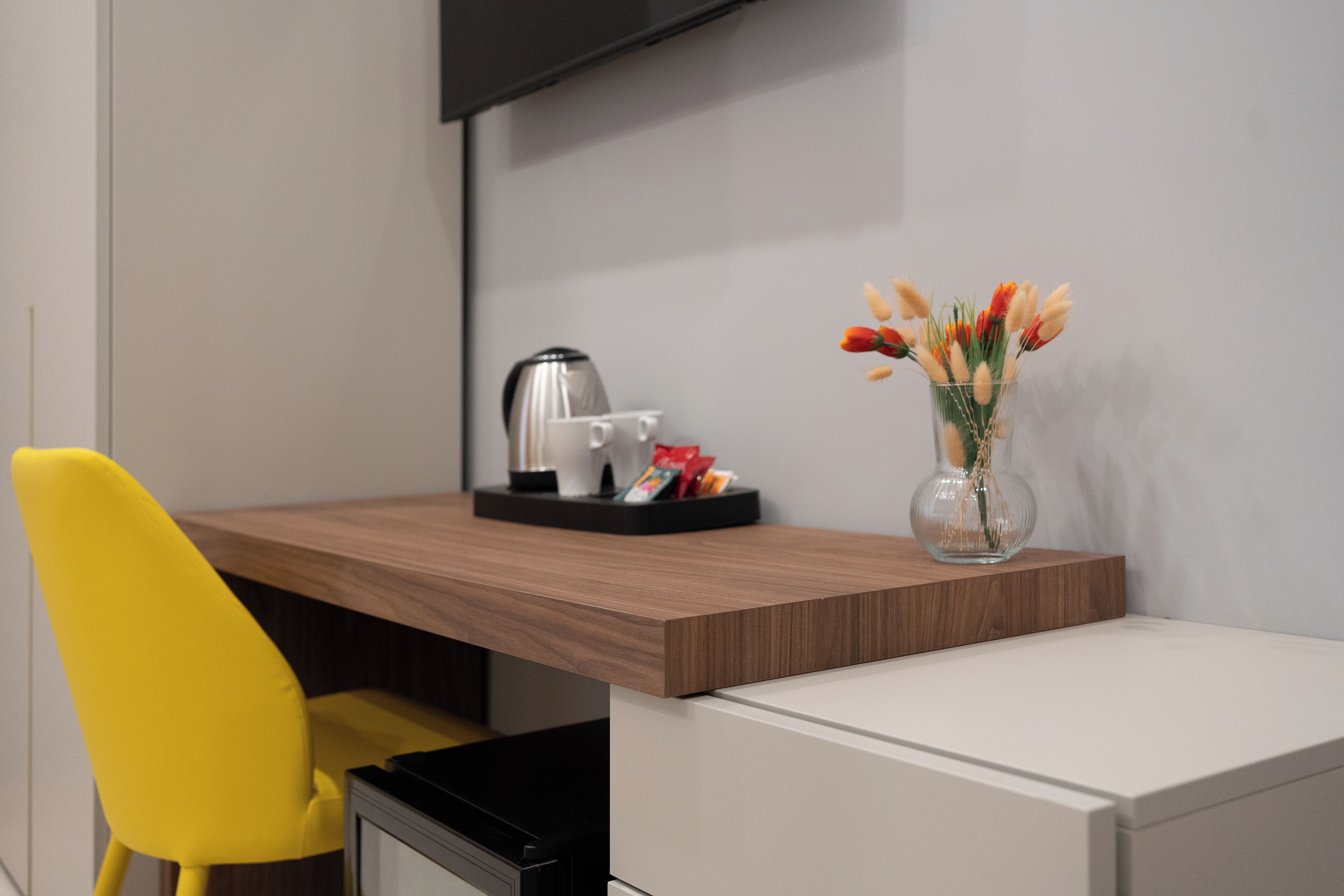 A modern hotel room with a wooden desk, a yellow chair, a white cabinet, and a vase with flowers.