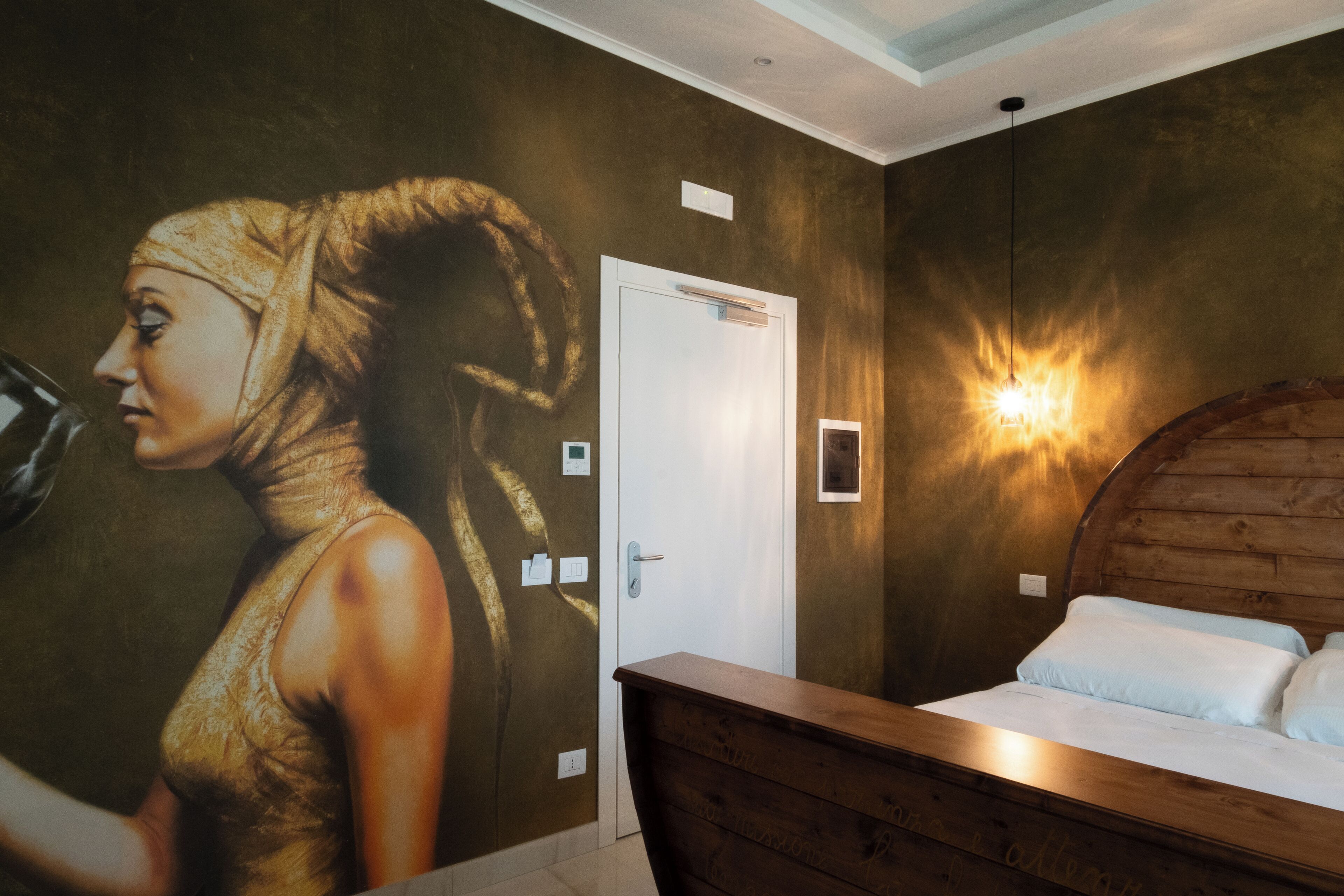 Una habitación con cama, un mural en la pared, una puerta y una lámpara fijada a la pared.
