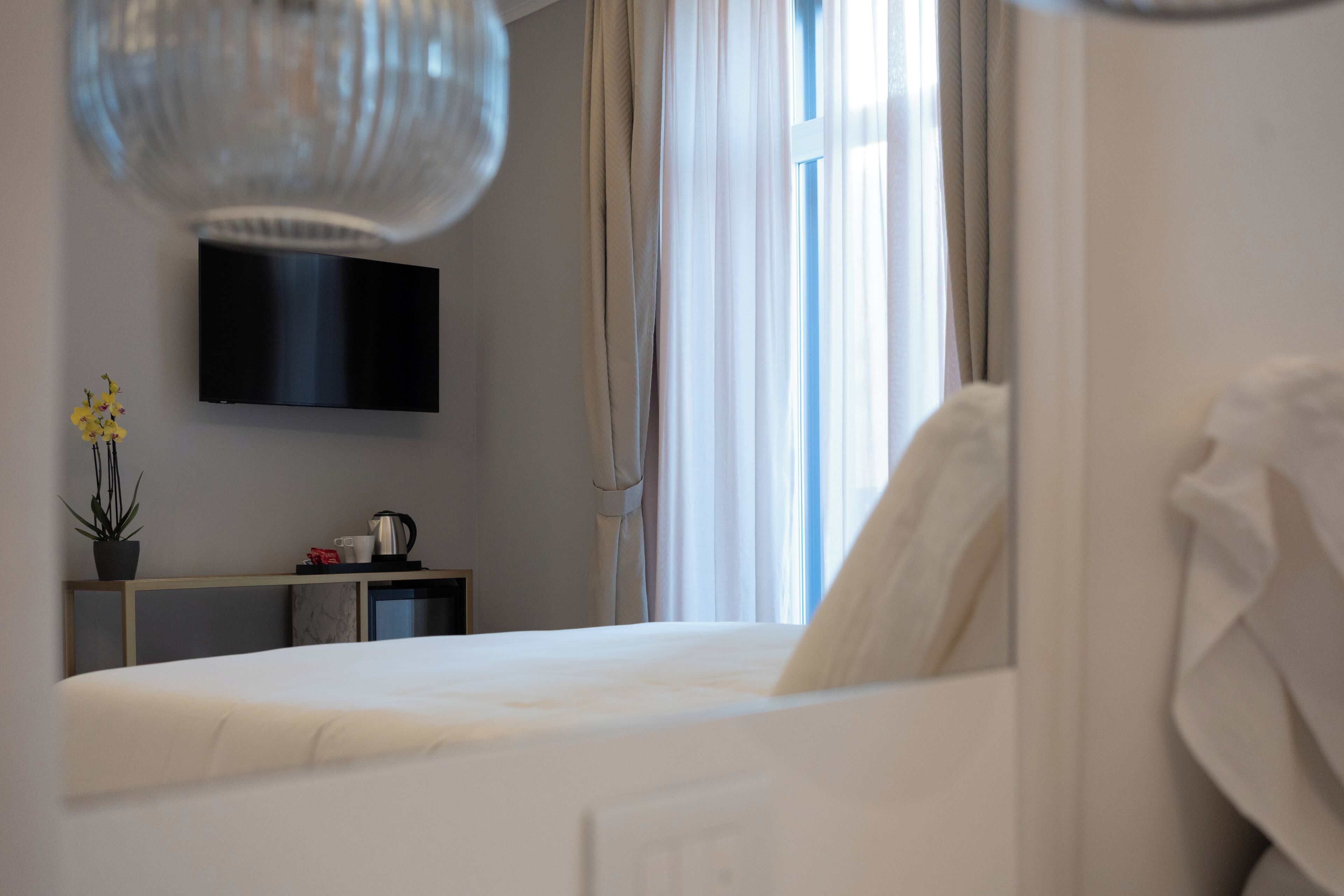 Une chambre d’hôtel comprenant un lit, un téléviseur à écran plat fixé au mur, une petite table avec une plante et une bouilloire, ainsi qu’une fenêtre avec des rideaux.