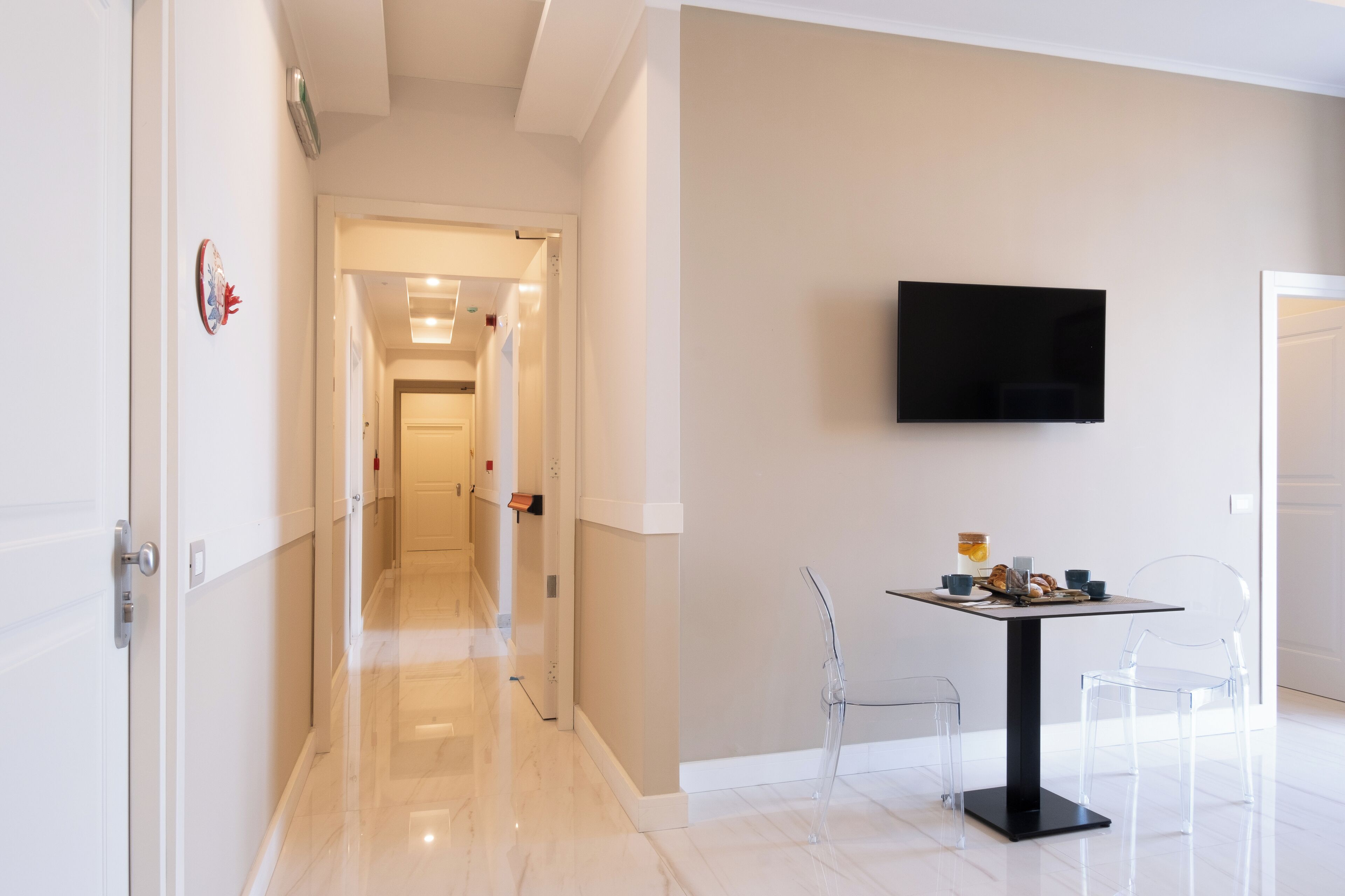 A modern hotel hallway with a dining area featuring a table and chairs, a mounted TV, and a clear view into the next room.
