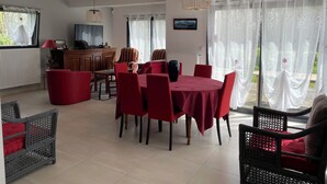 A dining area with a round table covered in a red tablecloth, surrounded by red chairs. There is a television on a wooden cabinet, and a window with sheer curtains. - Maison Récente Avec Tous les Équipements Traditionnels Très Bien Situé (Plouër-sur-Rance)