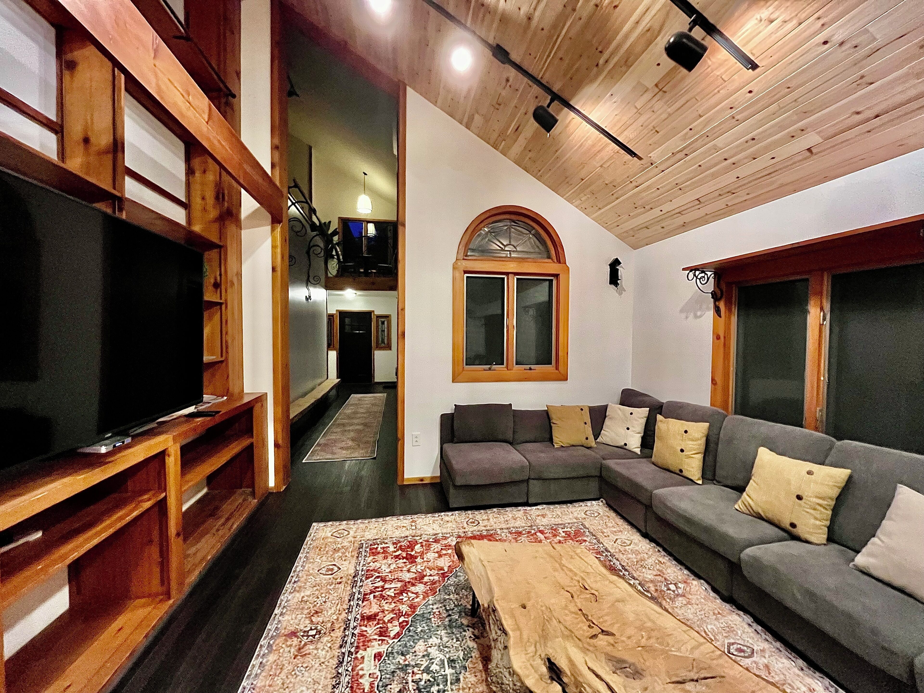 Un salon comprenant un téléviseur, une table basse en bois, un canapé gris et un tapis à motifs.