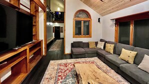 Un salon comprenant un téléviseur, une table basse en bois, un canapé gris et un tapis à motifs.