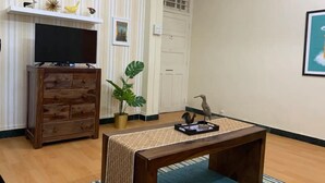 Un salon avec un meuble TV en bois, une plante, une table ornée d’un chemin de table tissé et un tableau encadré accroché au mur.