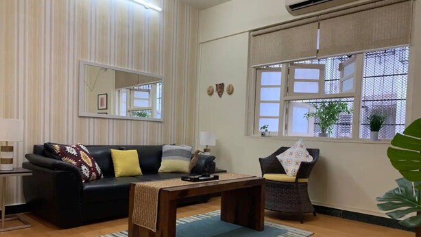 Un salon avec un canapé noir, une table basse en bois et un tapis à rayures. Il y a une fenêtre donnant sur l’extérieur et un miroir accroché au mur.