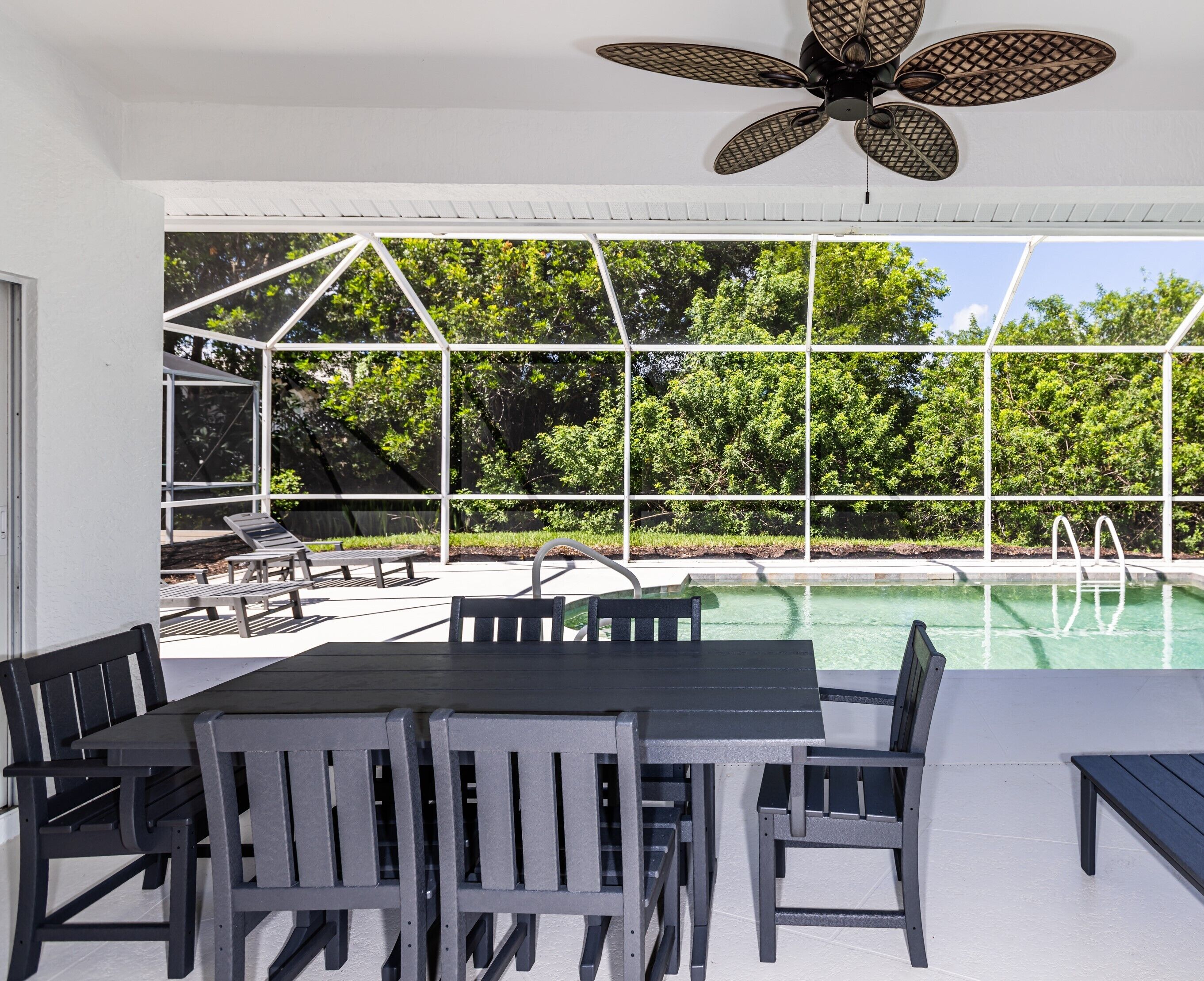 An outdoor pool area with a table and chairs, a ceiling fan, and a view of trees through large windows.