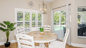 A dining area with a round table, white chairs, a potted plant, and a view of a pool through large windows.
