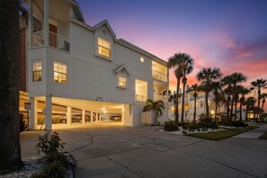 A multi-story hotel with a covered parking area, palm trees, and a sunset sky.