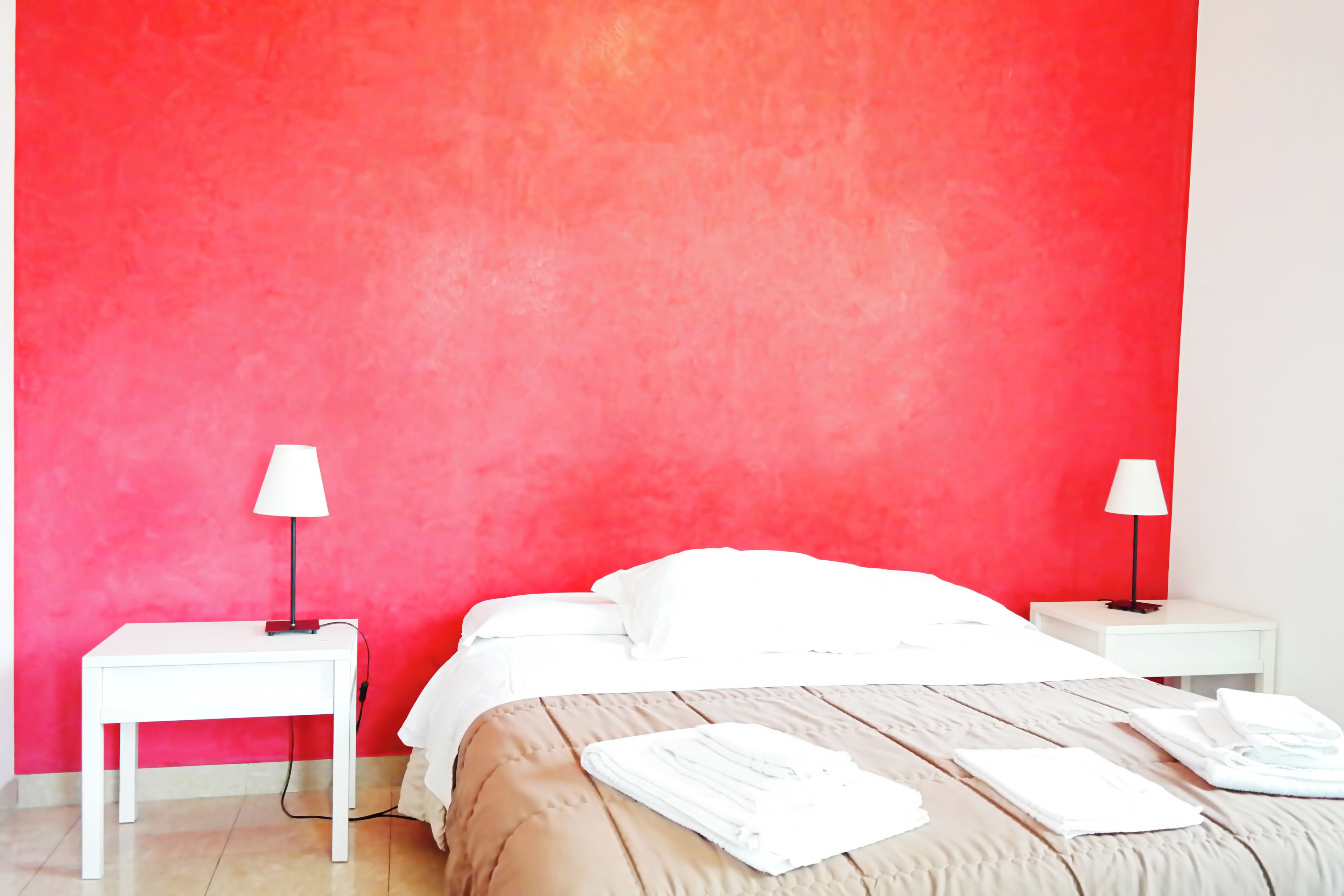 A hotel room with a red accent wall, a bed with white linens, two bedside tables with lamps, and a folded towel on the bed.