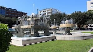 A public fountain with multiple water jets, surrounded by a well-maintained lawn and a few trees. - Harmony Central (Lecce)