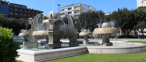 A public fountain with multiple water jets, surrounded by a well-maintained lawn and a few trees.