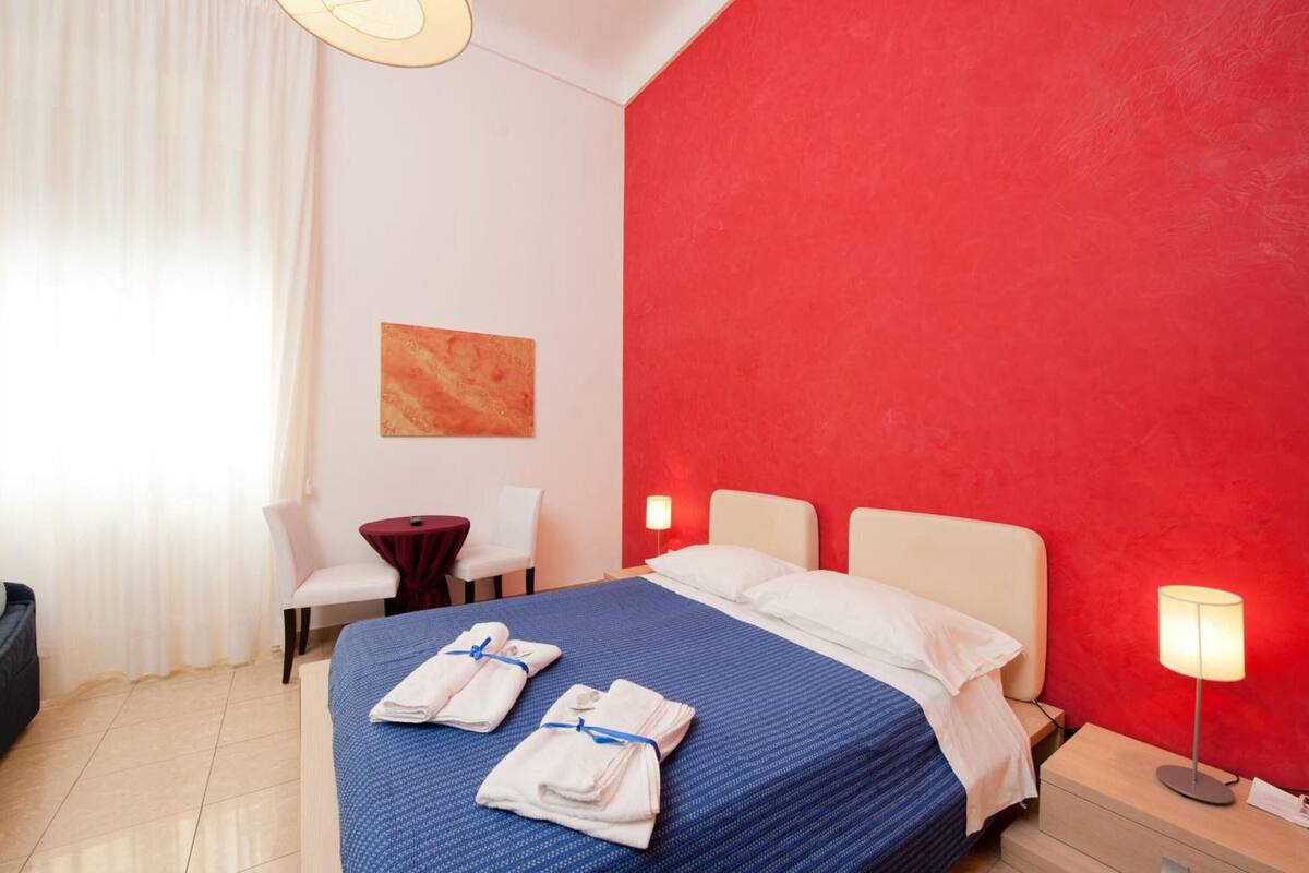 A hotel room with a bed, a desk, a chair, and a red accent wall.