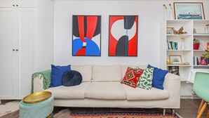 A living room with a beige sofa, colorful cushions, abstract wall art, a bookshelf, and a small table with a chair. - Dubrovnik art Gateway (Dubrovnik)